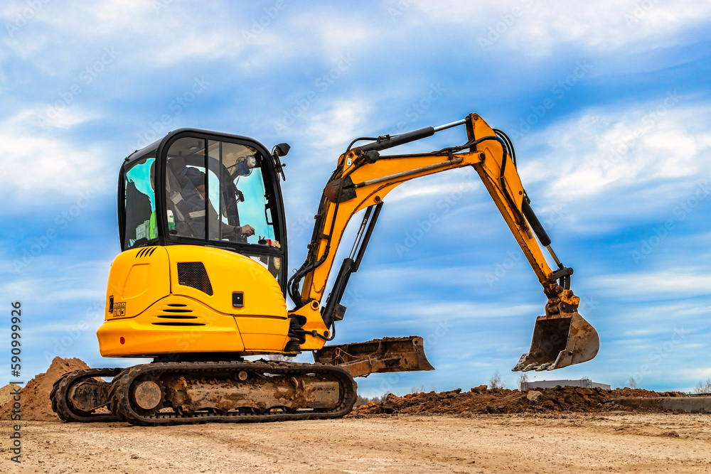 Mini excavator at the construction site on the edge of a pit against a cloudy blue sky. Compact construction equipment for earthworks. An indispensable assistant for earthworks. Mini excavator at the construction site on the edge of a pit against a cloudy blue sky. Compact construction equipment for earthworks. An indispensable assistant for earthworks.
