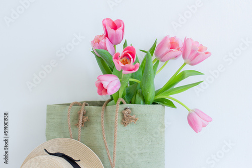 Pink tulips bouquet in green shopping bag and a straw hat on white background copy space