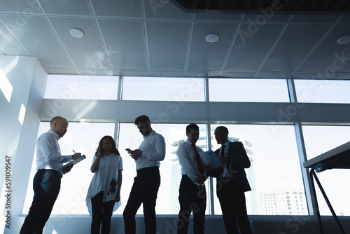 Silhouettes of people standing near a panoramic window in a modern office. Team of young professional business people working and chatting together in a meeting. © xartproduction