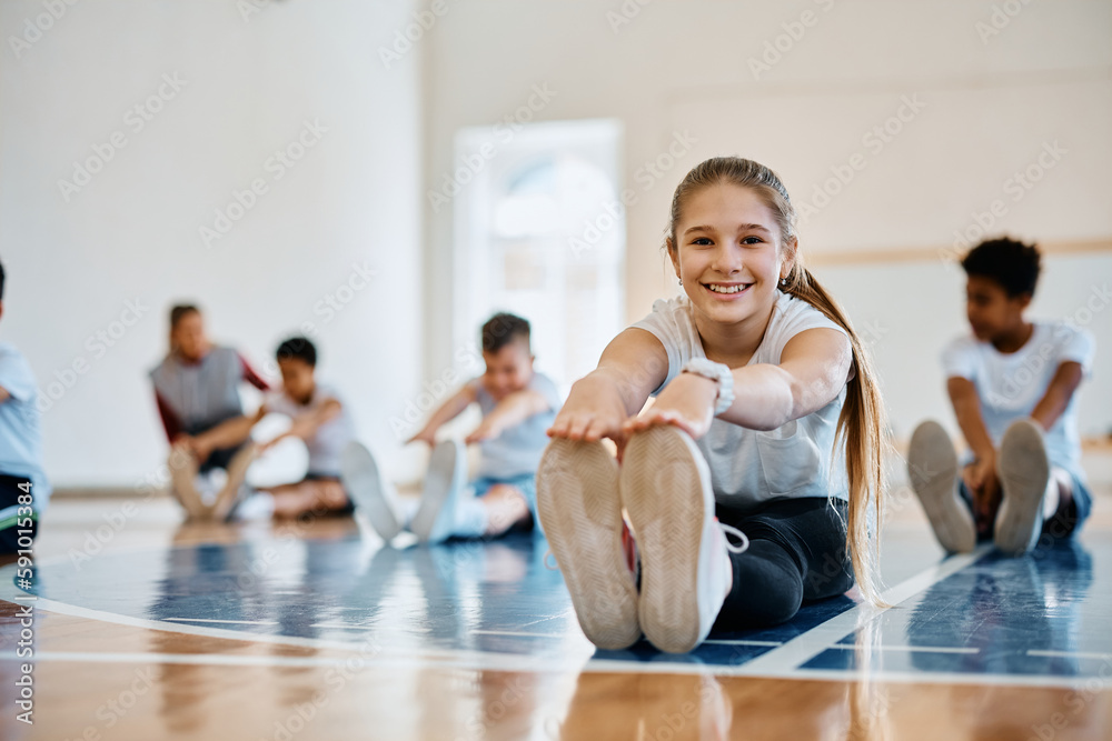 Happy schoolgirl stretching during PE class and looking ta camera. - obrazy, fototapety, plakaty 