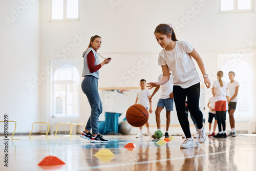 Fototapeta Naklejka Na Ścianę i Meble -  Schoolgirl practicing basketball while having PE class with sports teacher and classmates at school gym.