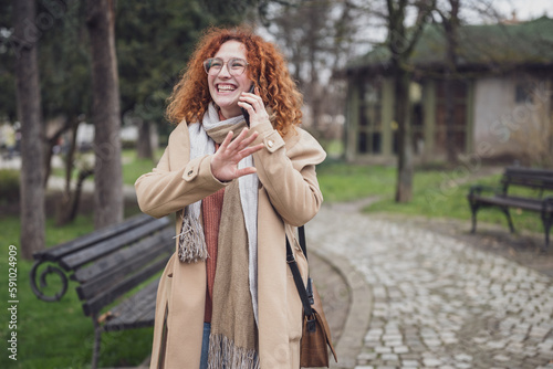 Natural portrait of smiling caucasian ginger woman with freckles and curly hair. She is talking on phone.