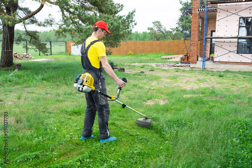 Male gardener mows the green grass of the lawn in the backyard at construction site with a gasoline mower. Trimmer for the care of a garden plot