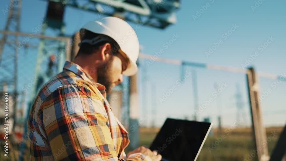 Serious attentive man engineer architect in protective helmet looks at tablet screen outdoors near power line in sunny day. Professional worker in power and energy industry works creates new project.