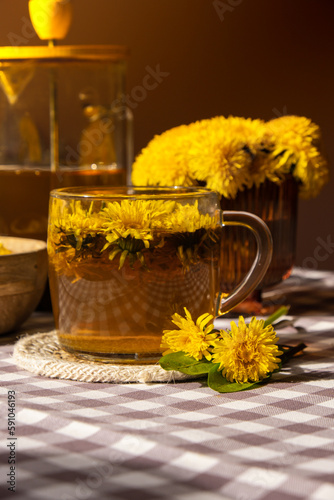Dandelion flower healthy tea in glass teapot and glass cup on table. Delicious herbal tea from fresh dandelion flowers at home at summer day. Green clearing Hot dandelion tea in a glass teapot