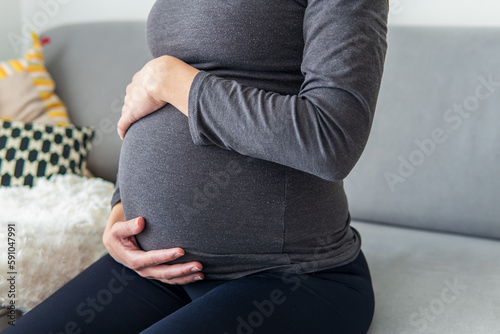 Pregnant woman sitting comfortably on the sofa and resting while putting her hands on her belly. Woman relaxing due to pregnancy pain and tiredness. Maternity, pregnancy concept. 