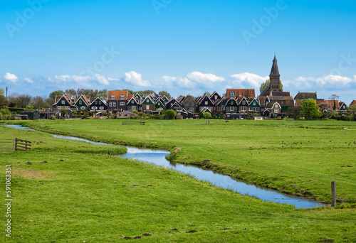 The quaint rural town of Marken island in the Netherlands is full of stunning architecture and nature, with a peaceful canal cutting through its grassy landscape beneath a blue sky. photo