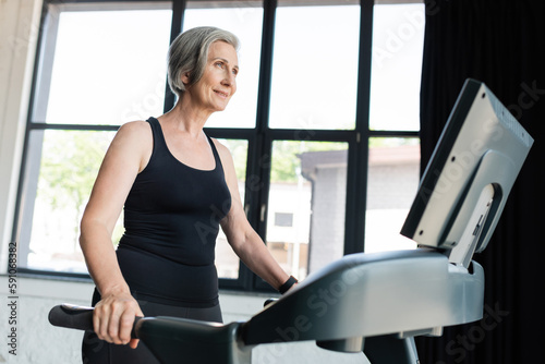 cheerful senior woman with grey hair running on treadmill in gym.