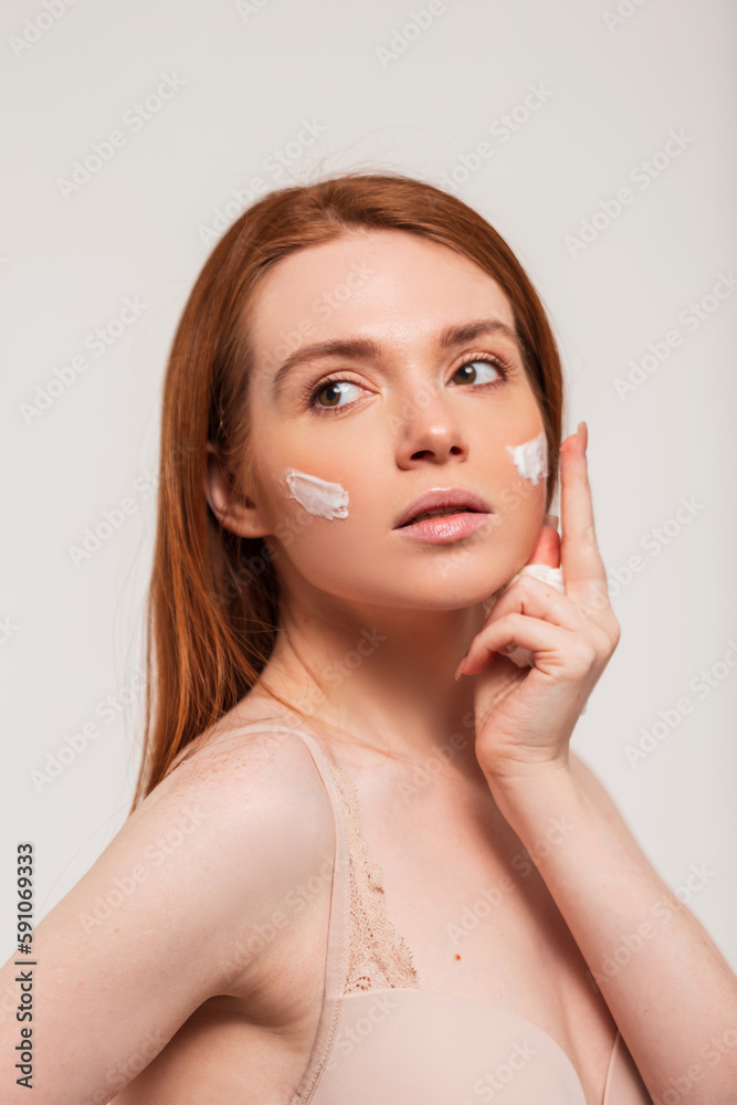 Red-haired beautiful fresh young woman applies cream on the skin of the face in the studio on a white background. Beauty and skin care Red-haired beautiful fresh young woman applies cream on the skin of the face in the studio on a white background. Beauty and skin care
