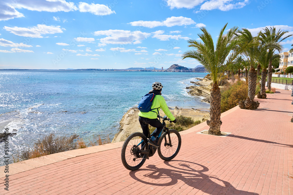 Fototapeta premium nice senior woman cycling on a cool spring day with her electric bicycle at the waterfront of Alicante, Costa Blanca, Spain