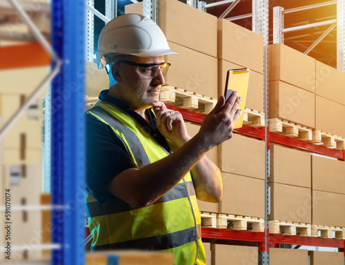 Customs warehouse worker. Man with tablet computer. Guy near racks with boxes. Customs warehouse worker thought for moment. Man in reflective vest and safety helmet. Career in bonded warehouse