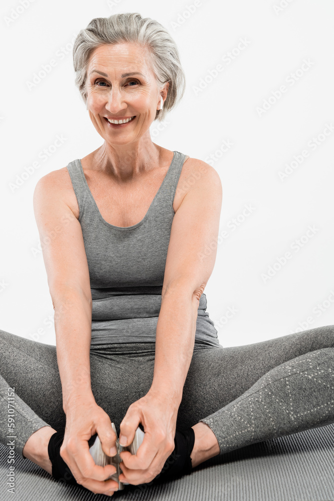 positive senior woman with wireless earphones sitting in yoga pose on mat isolated grey.