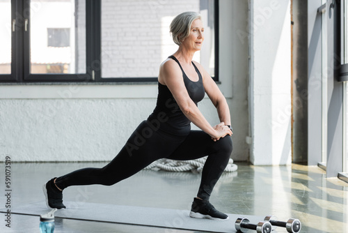 full length of senior woman in sportswear doing lunges on fitness mat near dumbbells in gym.