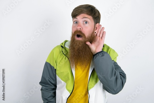 Oh my God! Funny astonished red haired man wearing sportswear standing over white studio background opening mouth widely looking aside, with hand near ear trying to listen to gossips.