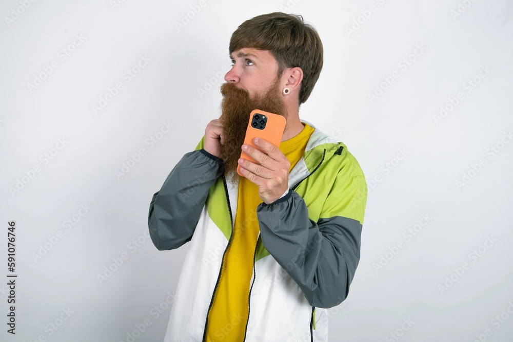 Thinking dreaming red haired man wearing sportswear standing over white studio background using mobile phone and holding hand on face. Taking decisions and social media concept. Thinking dreaming red haired man wearing sportswear standing over white studio background using mobile phone and holding hand on face. Taking decisions and social media concept.