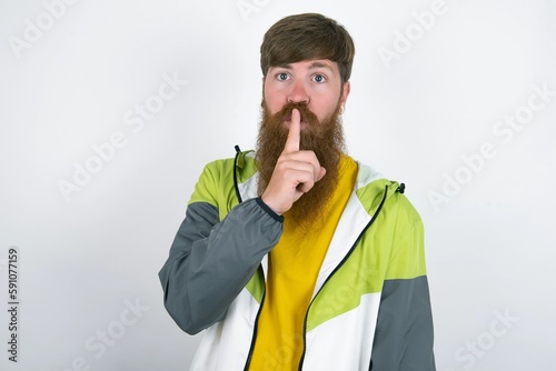 Surprised red haired man wearing sportswear standing over white studio background makes silence gesture, keeps finger over lips and looks mysterious at camera photo