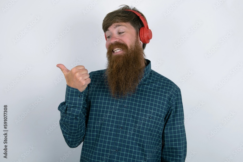 Fototapeta premium red haired man wearing plaid shirt over white studio background listens audio track via wireless headphones points thumb away advertises copy space