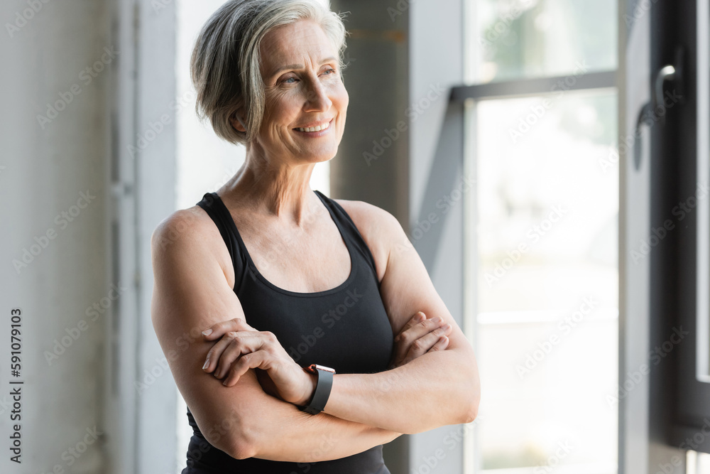 happy senior sportswoman in tank top smiling while standing with crossed arms in gym.