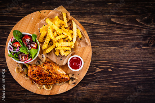 Roast chicken breast, French fries, fried mushrooms and fresh vegetables on wooden table

