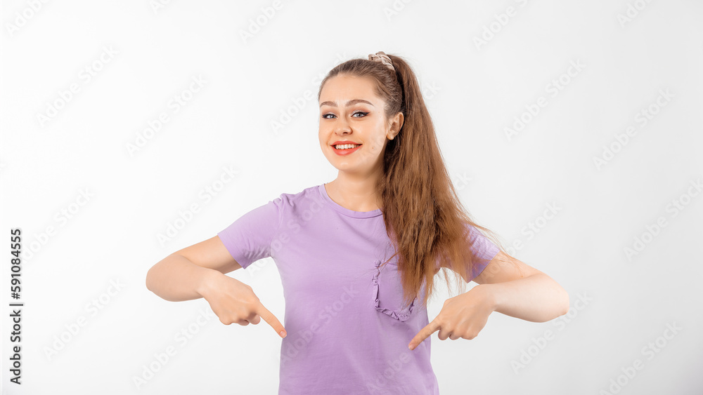 Happy young woman with long hair pointing fingers down and smiling, showing advertisement, product below, promo offer, standing in casual t shirt over white background Happy young woman with long hair pointing fingers down and smiling, showing advertisement, product below, promo offer, standing in casual t shirt over white background