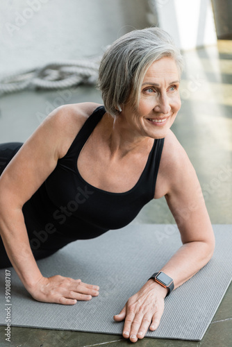 joyful senior sportswoman smiling while exercising on fitness mat in gym.