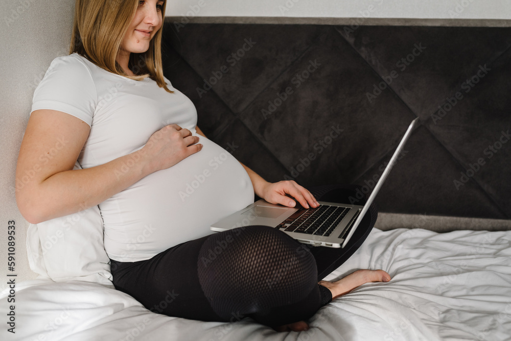 Naklejka premium Happy pregnant student girl studying at home, learning webinar, workshop, teachers tutorial, writing notes on laptop. Expecting mom working and making video call. Using computer to watch movie online.