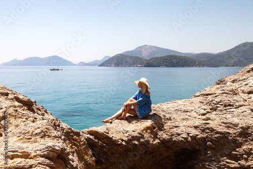 Lovely young woman in blue beachwear and hat sitting on rock at tropical azure ocean background, summer vacation. Charming tourist lady enjoy rest on sea. Travel holidays concept. Copy ad text space