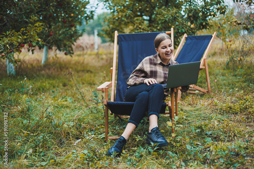 Stylish woman sitting on a chair near green plants in the garden and browsing the internet on a laptop while looking to the side. Remote work  female freelancer