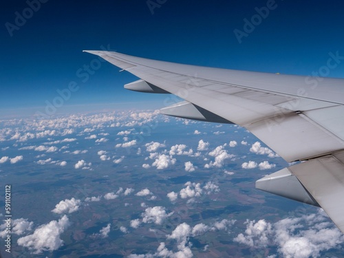 Wing of an airplane in blue sky with white fluffy clouds flying above green fields