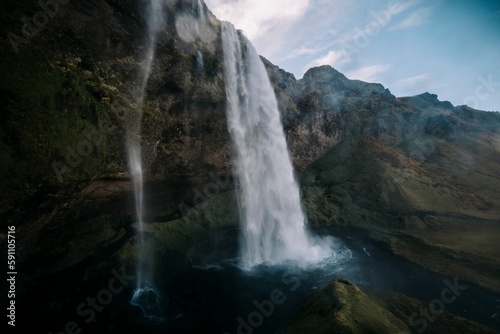Scenic view of a waterfall in green mountains on a cloudy day in Iceland © Rachelmcgrath/Wirestock Creators