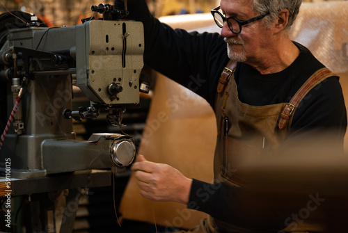 adult craftsman sitting adding sewing thread in a sewing machine to start sewing parts of a chair made of leather in his workshop, downtown rome, italy
