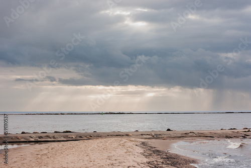 seashore with a sand embankment leading to the jetty. dramatic sky with clouds through which the rays of the sun shine.