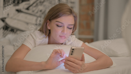 Young Woman using Smartphone while Lying in Bed on Stomach
