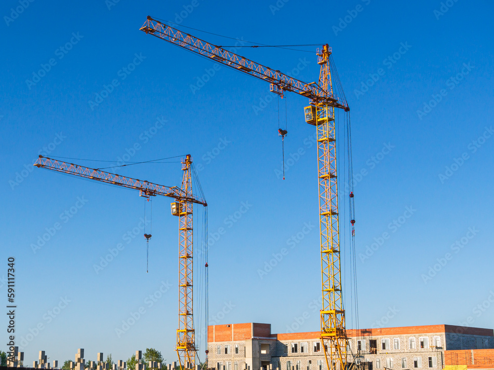 Construction of buildings outside the city. Equipment for construction and lifting of large loads with construction materials. Yellow cranes and blue sky