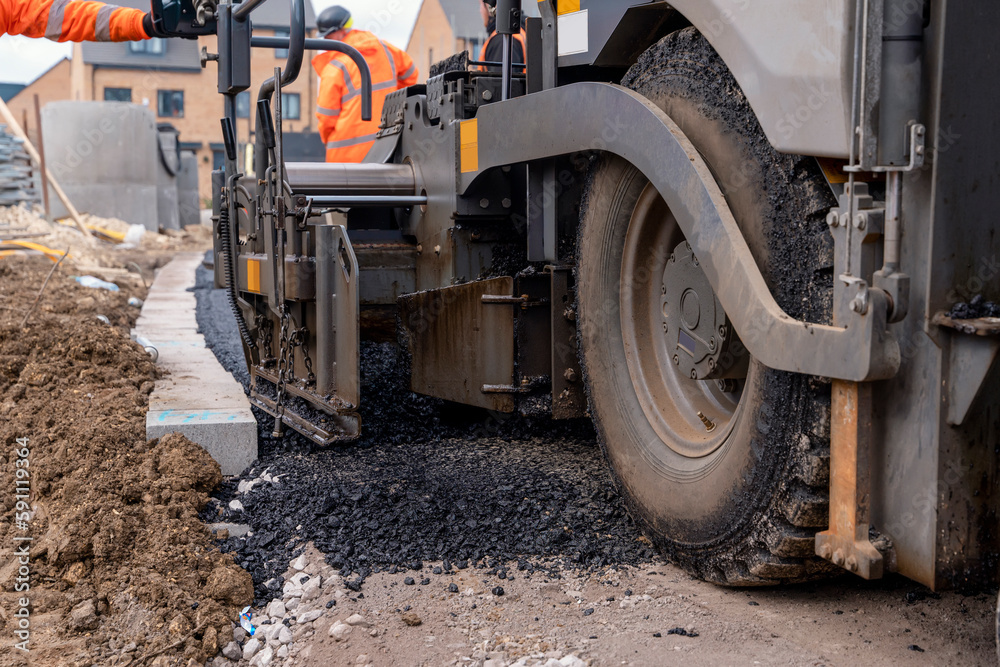 Asphalt paver filled with hot tarmac laying new road surface on new residential housing development site and roadworker operator in orange hi-viz next to it Asphalt paver filled with hot tarmac laying new road surface on new residential housing development site and roadworker operator in orange hi-viz next to it