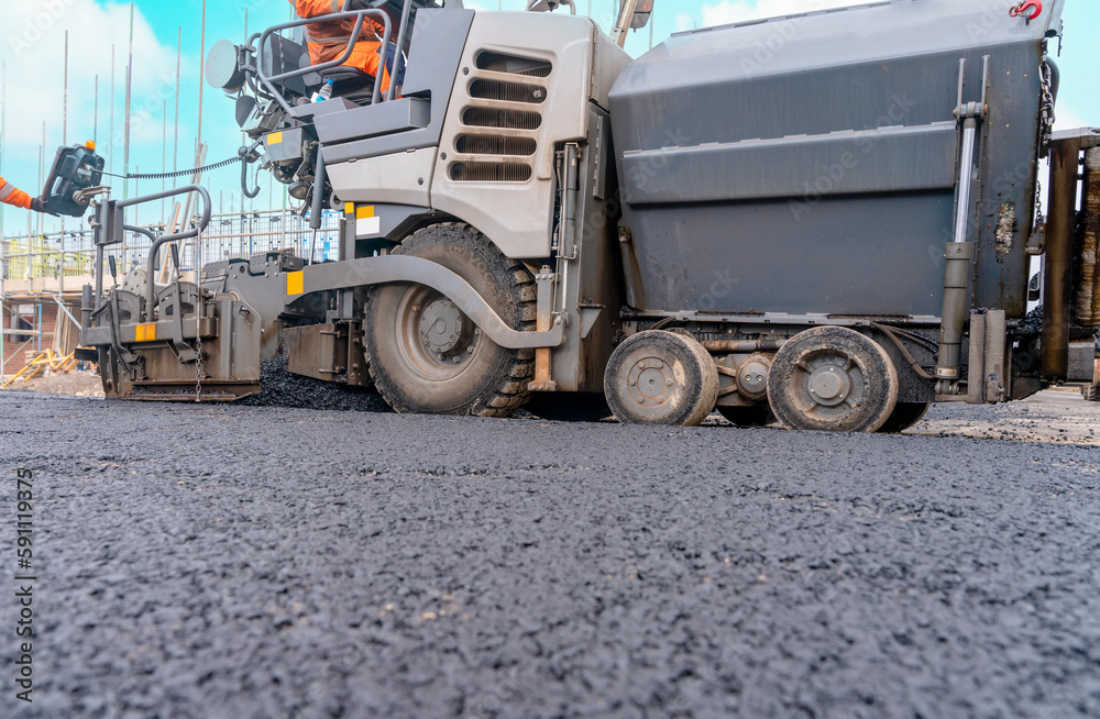 Asphalt paver filled with hot tarmac laying new road surface on new residential housing development site and roadworker operator in orange hi-viz next to it Asphalt paver filled with hot tarmac laying new road surface on new residential housing development site and roadworker operator in orange hi-viz next to it