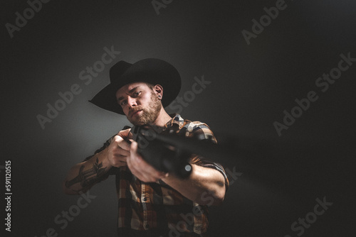Rugged man with beard and a shotgun in cowboy hat. Plaid shirt. Blue jeans. Studio session.