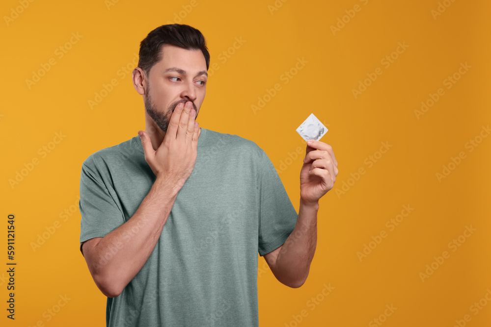 Confused man holding condom on orange background. Space for text Confused man holding condom on orange background. Space for text