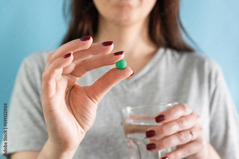 Woman taking green headache pill or painkiller drug with glass of water on blue background. Taking medicine