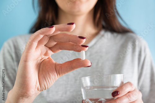 Woman taking white pill of statin medicine to treat high cholesterol or painkiller with glass of water on blue background photo