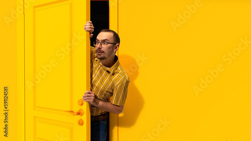Young man in glasses peeking out yellow door and looking with excitement, shock and positive impression. Big sales, success, win. Concept of emotions, facial expression, lifestyle, sales photo