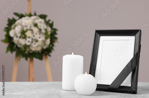 Bild auf Leinwand Photo frame with black ribbon, burning candles on table and wreath of plastic flowers near grey wall, space for text