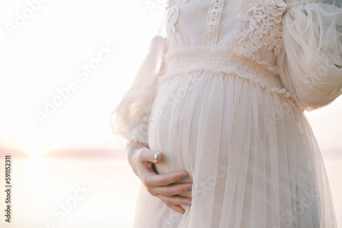 a pregnant girl supports her belly with her hand, a beautiful airy dress at sunset