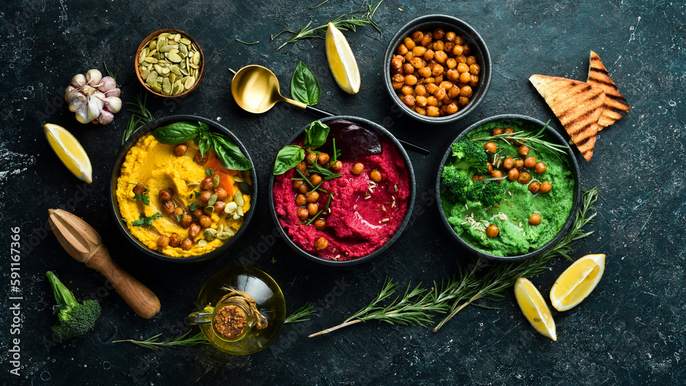 Colorful hummus bowls - green, yellow and beetroot hummus on dark background with lemon, olive oil, and spices. On a concrete black background.