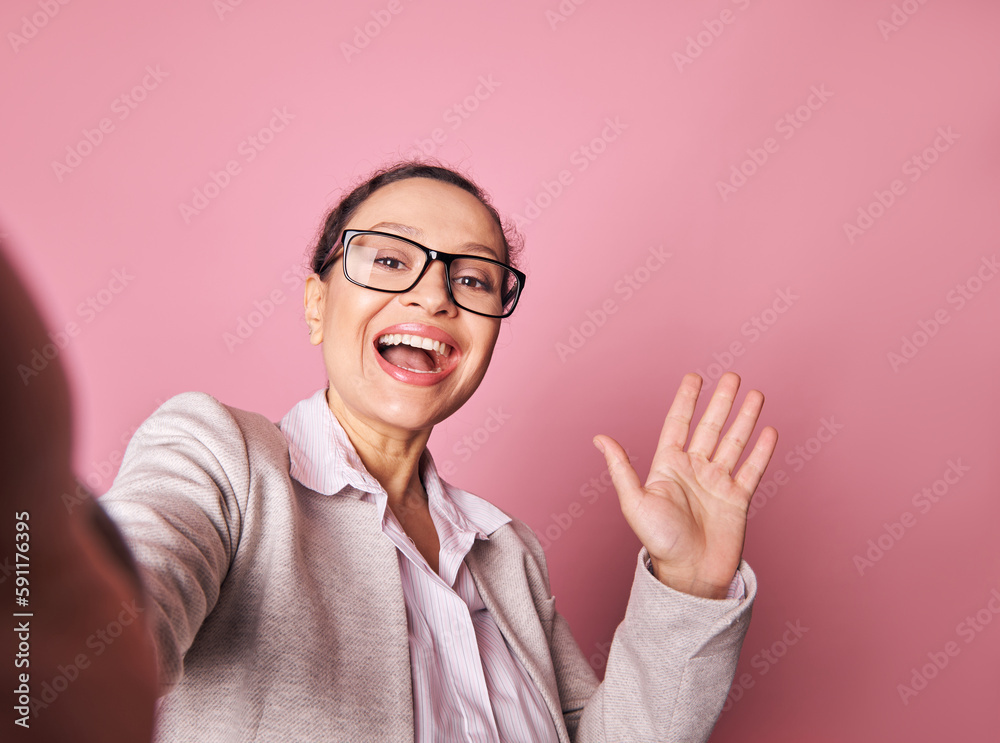 Selfie of stunning Latin American beautiful woman in gray jacket, smiling and greeting with her hand. Pretty brunette holds mobile phone, makes a self-portrait on her smartphone over pink background Selfie of stunning Latin American beautiful woman in gray jacket, smiling and greeting with her hand. Pretty brunette holds mobile phone, makes a self-portrait on her smartphone over pink background