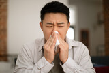 Portrait of ill middle aged asian man sitting at home, sneezing and using napkin, suffering from cold, flu or allergy