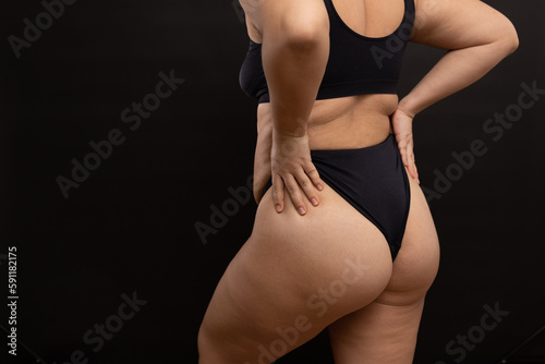 Overweight woman in black underwear pull up panties, back view. Flaunt figure imperfections, cellulite hips. Studio portrait over black background. Concept of obesity, body positive, self acceptance.