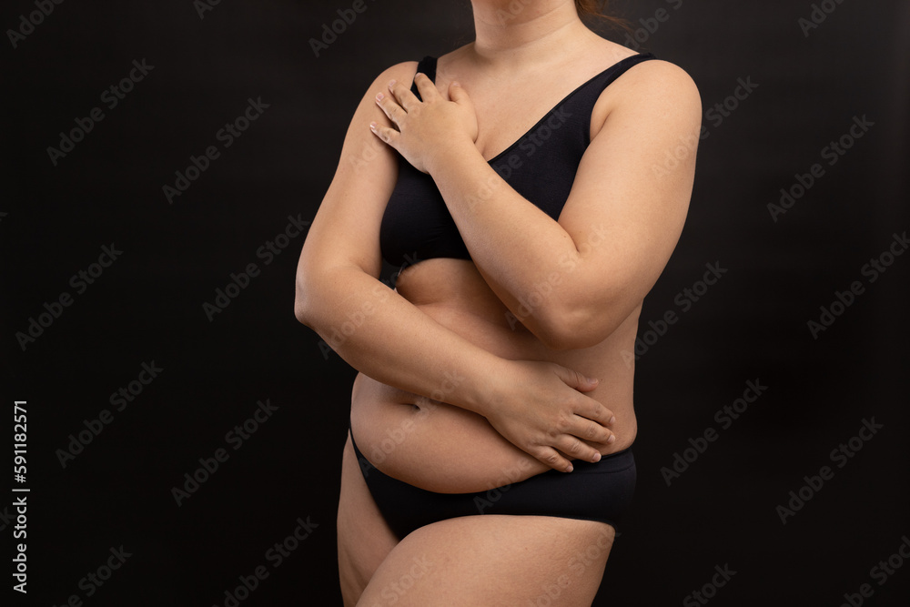 Naklejka premium Plump woman in black underwear covering with hands naked body parts. Hide figure imperfections, fat body. Studio portrait over black background. Concept of obesity, body positive, plus size people.