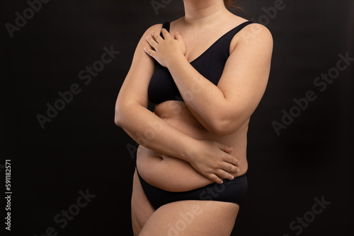 Plump woman in black underwear covering with hands naked body parts. Hide figure imperfections, fat body. Studio portrait over black background. Concept of obesity, body positive, plus size people.