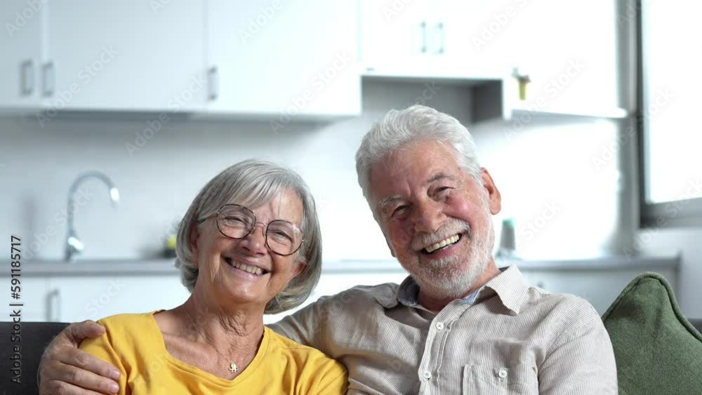 Headshot portrait of smiling elderly 60s husband and wife sit relax on couch hugging cuddling, happy mature old couple rest on sofa in living room embrace look at camera show love and care
